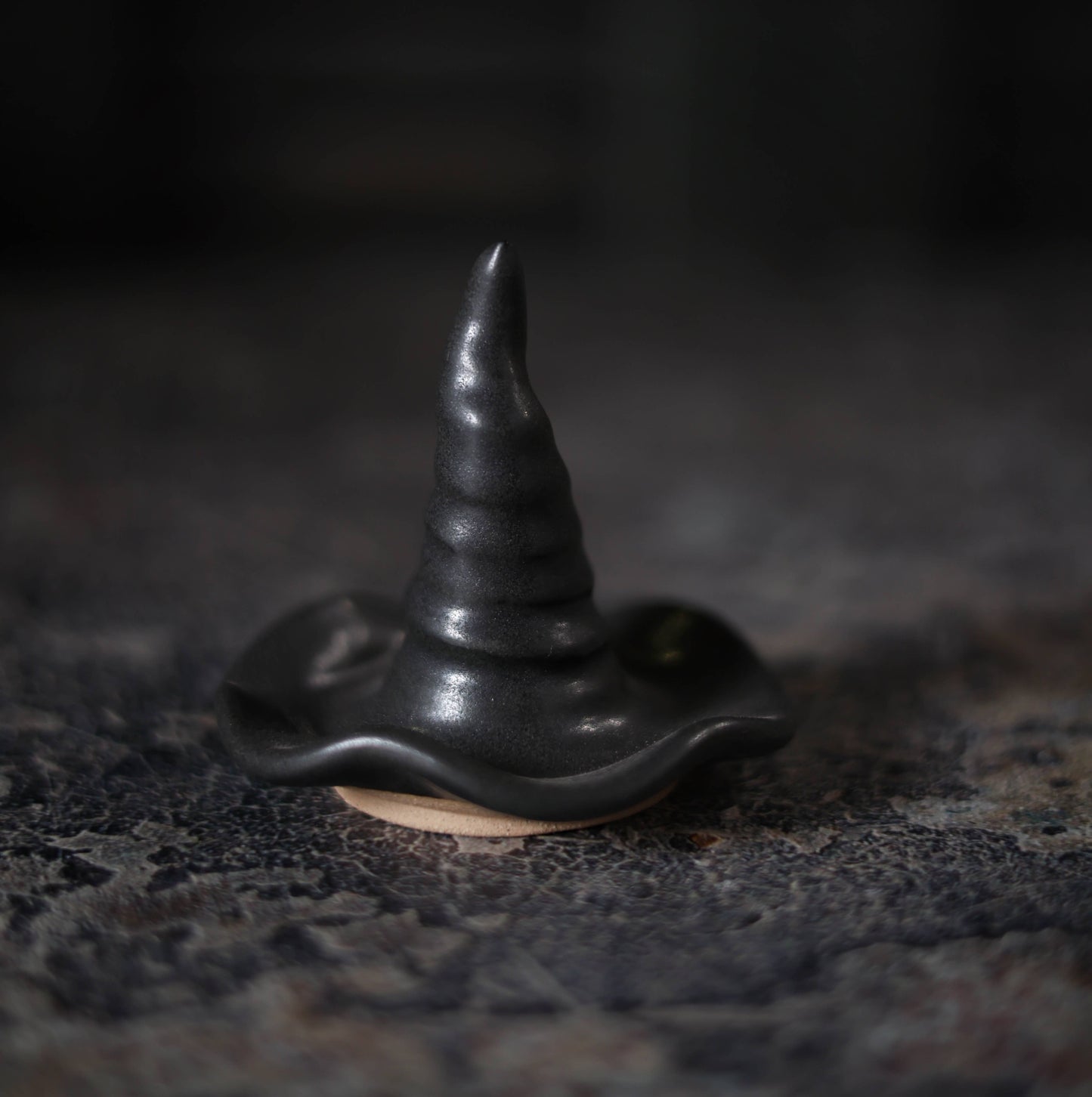 A small black ceramic witch hat incense holder with a pointed conical top and wavy brim, resting on a dark textured surface.