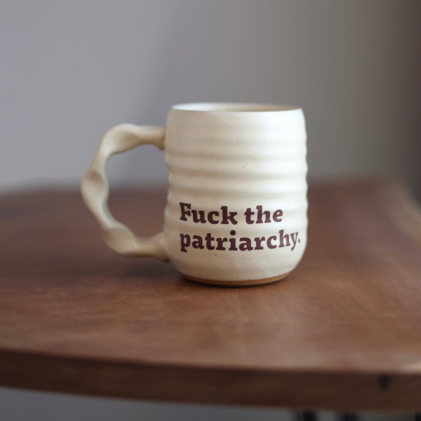 White ceramic mug with 'Fuck the patriarchy.' text on a wooden surface.