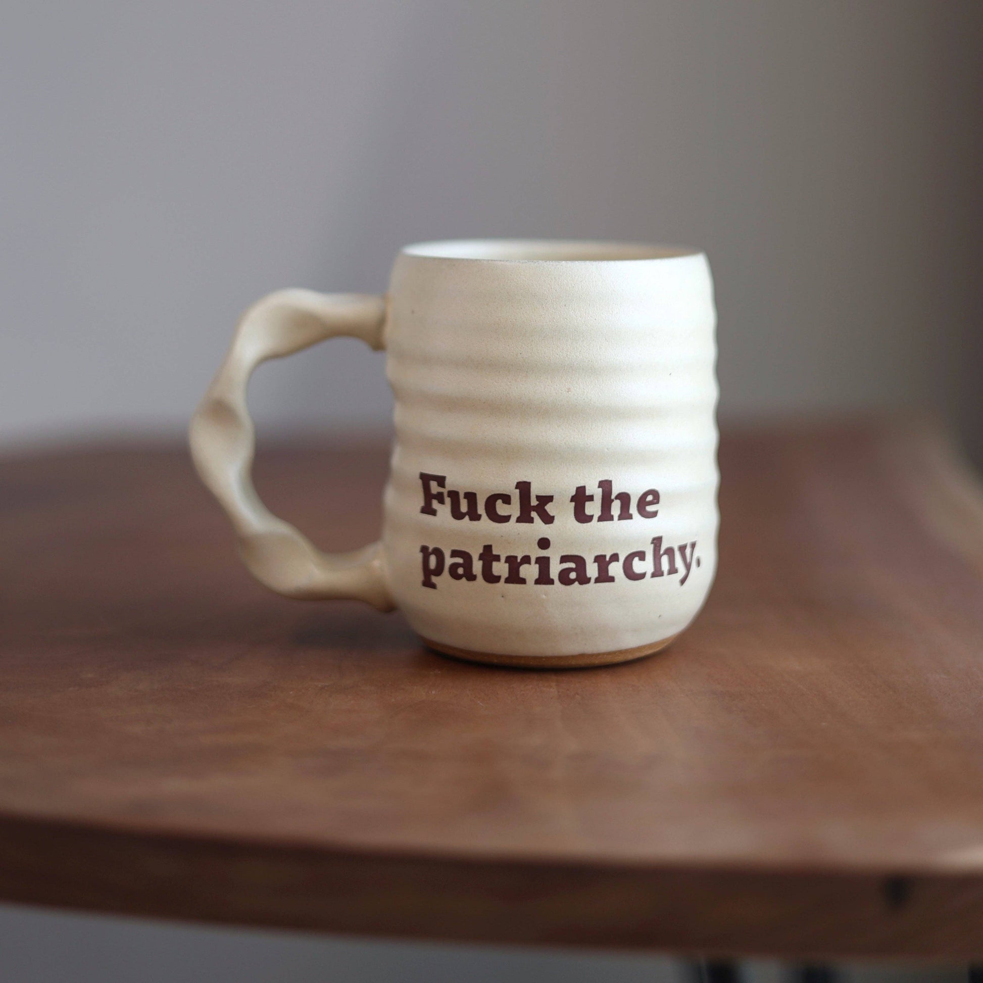 White ceramic mug with 'Fuck the patriarchy.' text on a wooden surface.