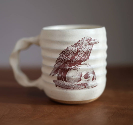 An off-white ceramic mug with a ribbed texture, featuring a sepia illustration of a raven perched on a skull. The mug is shown on a wooden surface with a blurred gray background.