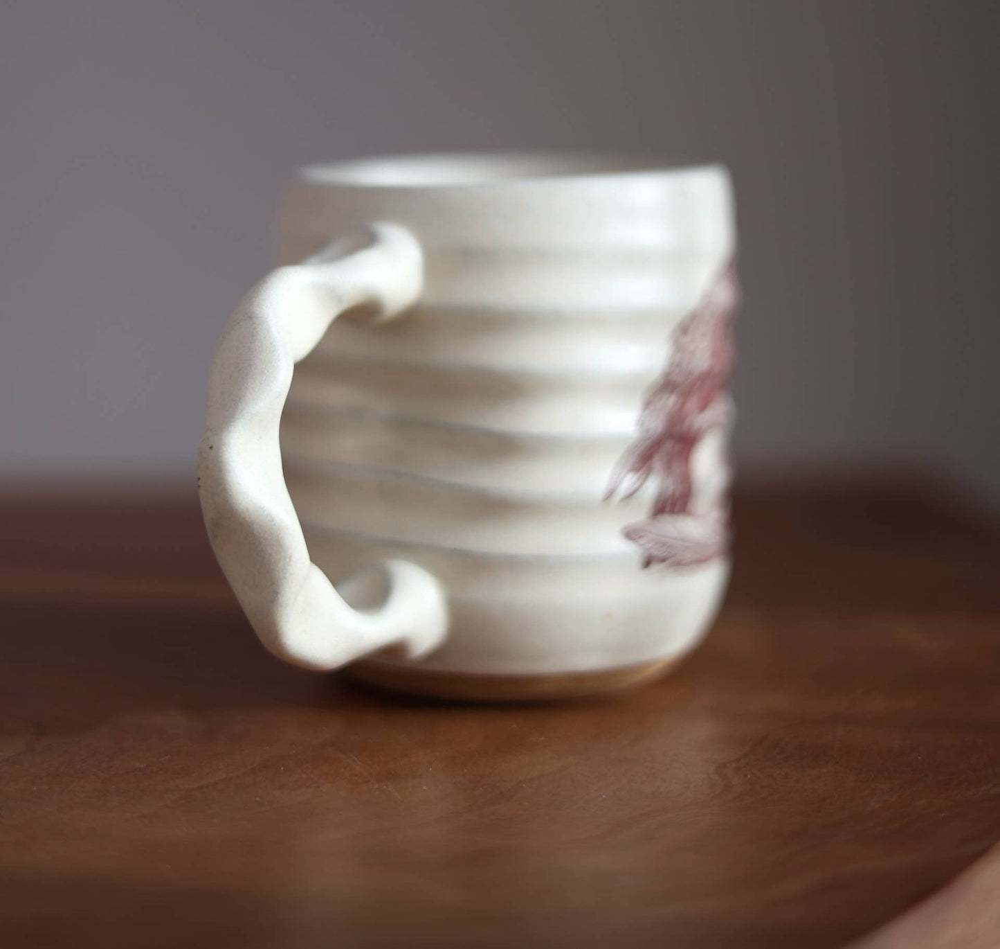 A handmade ceramic mug with a textured, ribbed body and a sculptural handle, shown sitting on a wooden surface against a neutral gray background.