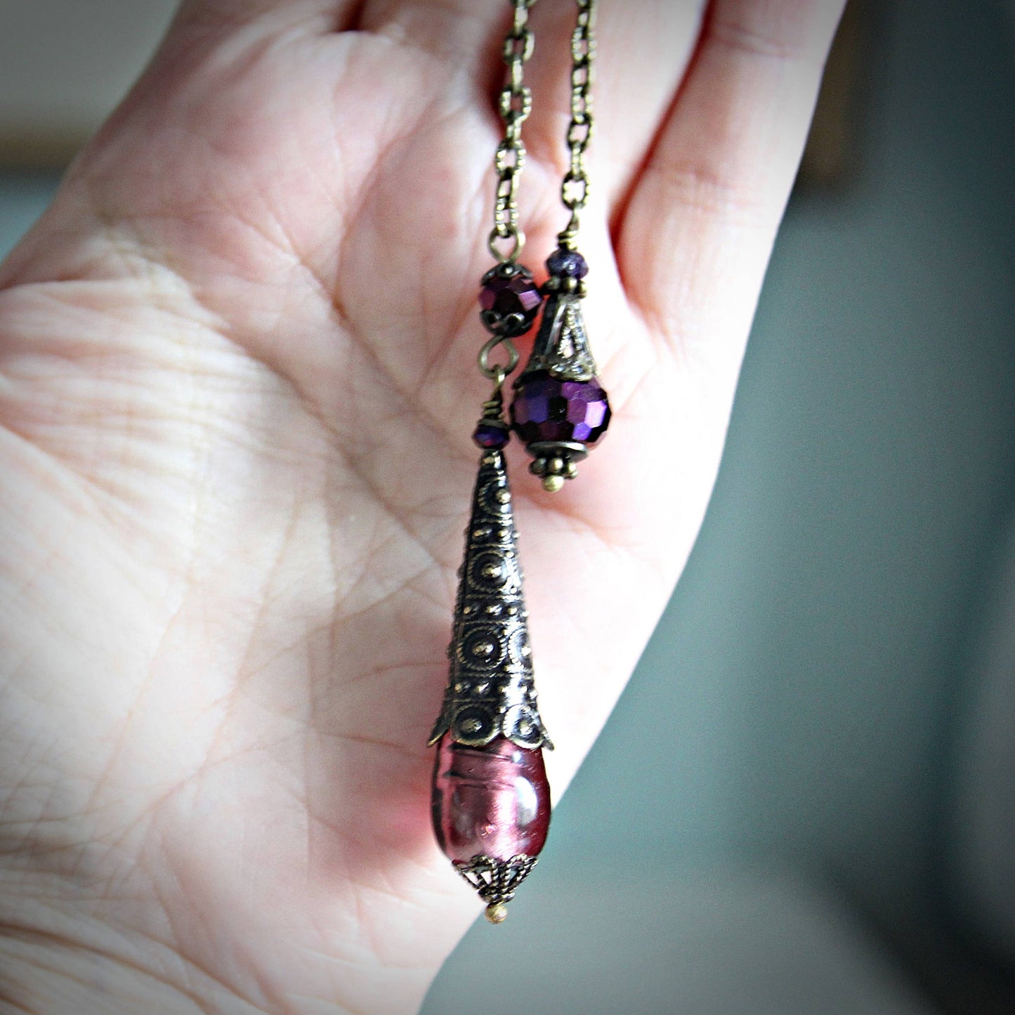 Pendulum with purple teardrop pendant held in a hand against a neutral background