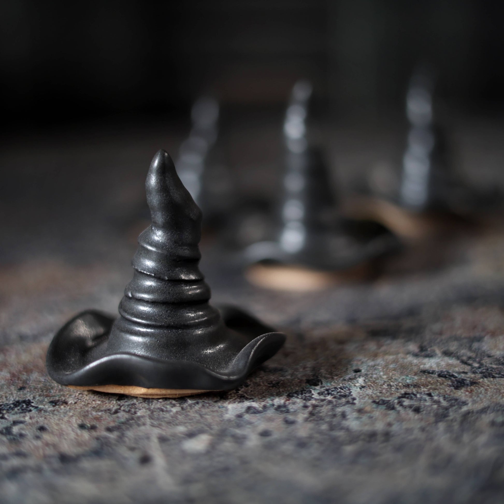 A small black witch hat-shaped object with a pointed top and wavy brim, photographed close up on a textured dark surface with blurred matching objects in the background.