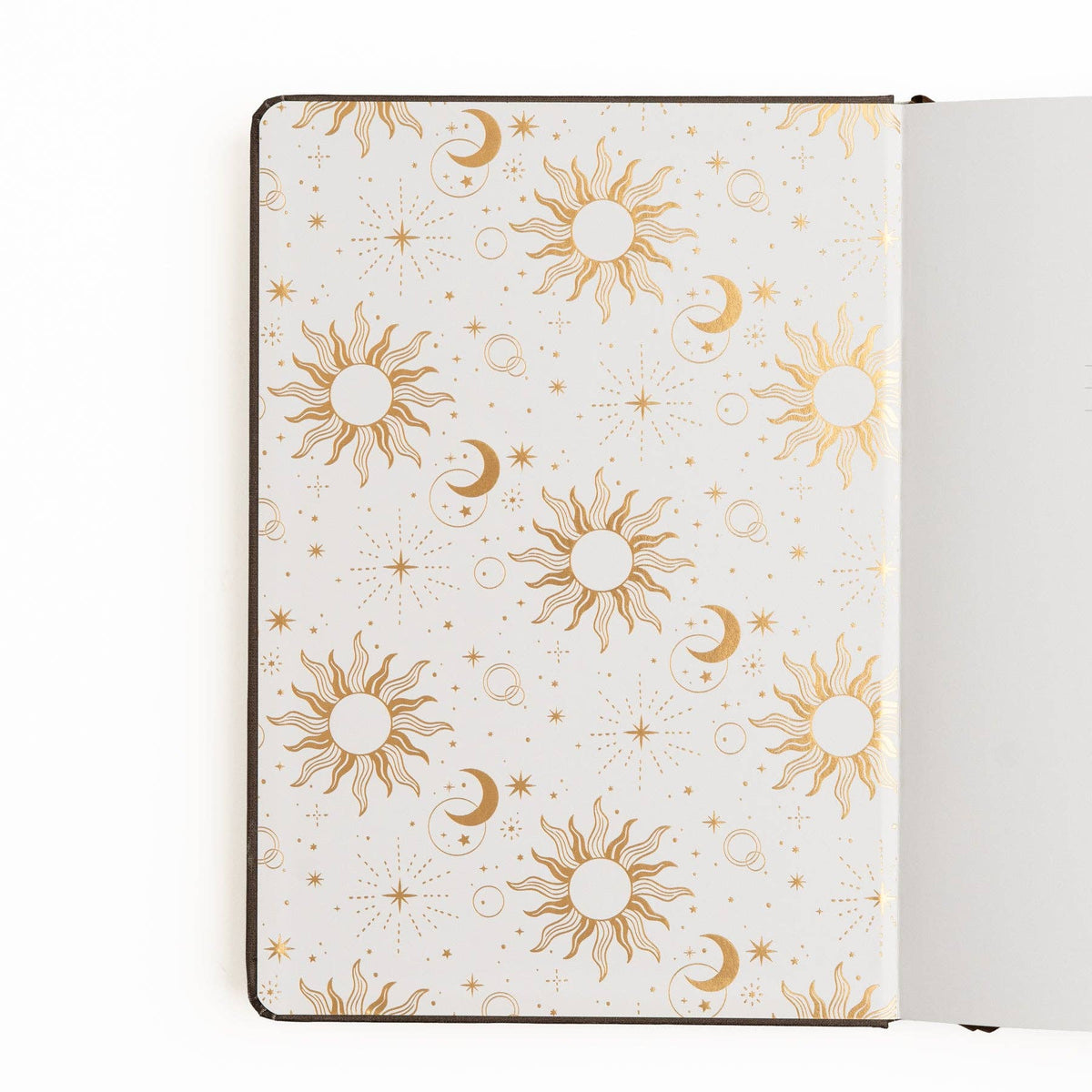 A notebook or journal shown open from above, with the left page featuring a repeating celestial pattern of gold suns, crescent moons, stars, and dots on a warm off-white background. The right page is blank and white.