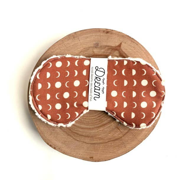 A fabric sleep sack with a rust-orange and cream moon phase pattern, displayed on a round wooden slice against a white background. A white paper band with the brand name 'Dream' is wrapped around the center.