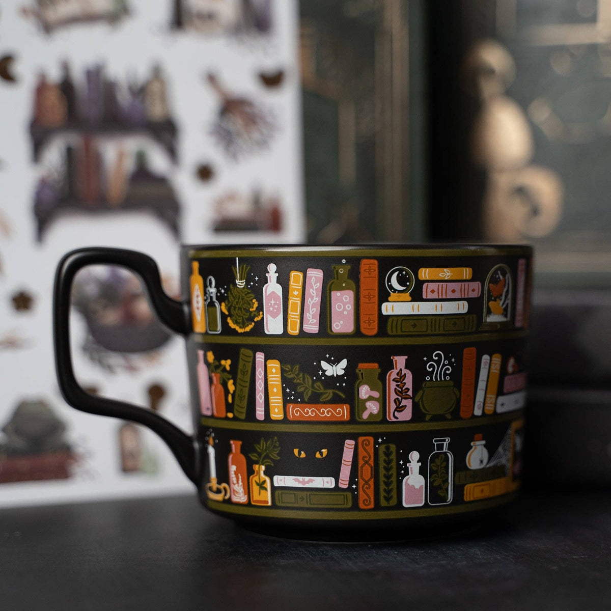 A black ceramic mug with a detailed illustration of occult bookshelves, potion bottles, and magical symbols, set against a dark, moody background with a blurred bookshelf and mystical figurine.