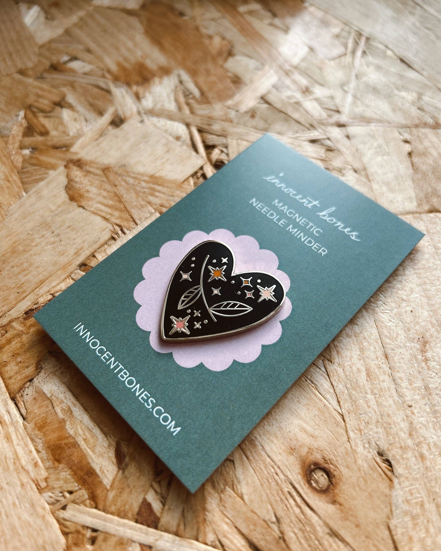 Heart-shaped pin with star and moon design on a green card with 'Innocent Bones' branding, placed on wooden surface.