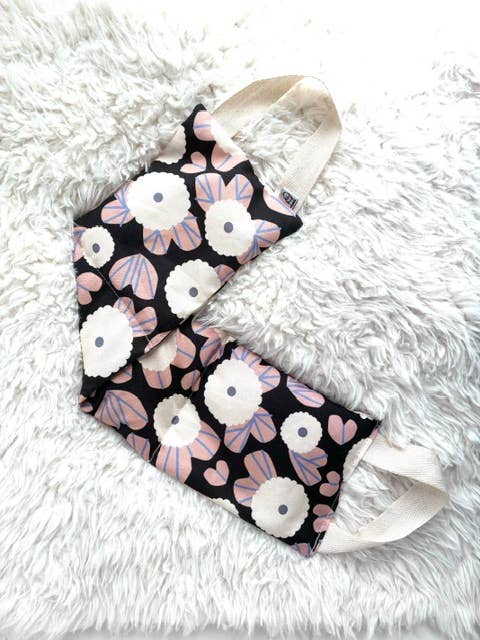 Two reusable cloth face masks with a black background and a floral pattern of white and pink flowers, laid flat on a white faux fur surface.