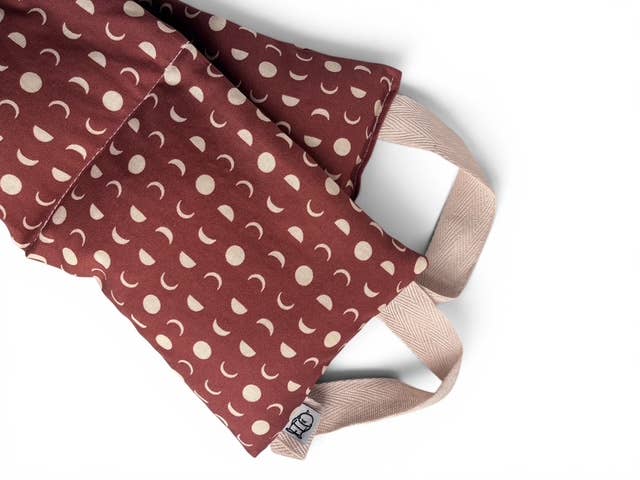 A close-up of a fabric face mask with a dusty rose red background and a repeating pattern of cream-colored crescent moons and dots. The mask features light beige adjustable straps and a small fabric brand tag reading 'hcl'.