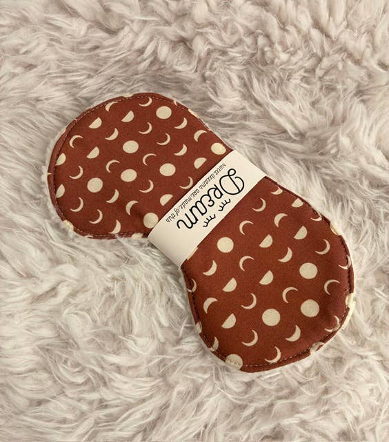 A reusable menstrual cup with a brown and cream moon phase pattern, wrapped in a white paper band that reads 'Dream' and 'reusable menstrual cup', resting on a white faux fur surface.
