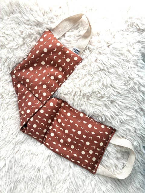 A reusable cloth face mask with a rust-brown and cream crescent moon and dot pattern, laid flat on a white faux fur surface.