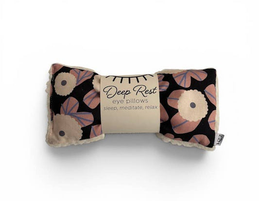 A rolled eye pillow with a floral pattern in pink, beige, and black, wrapped in a paper band that reads 'Deep Rest eye pillows sleep, meditate, relax'.