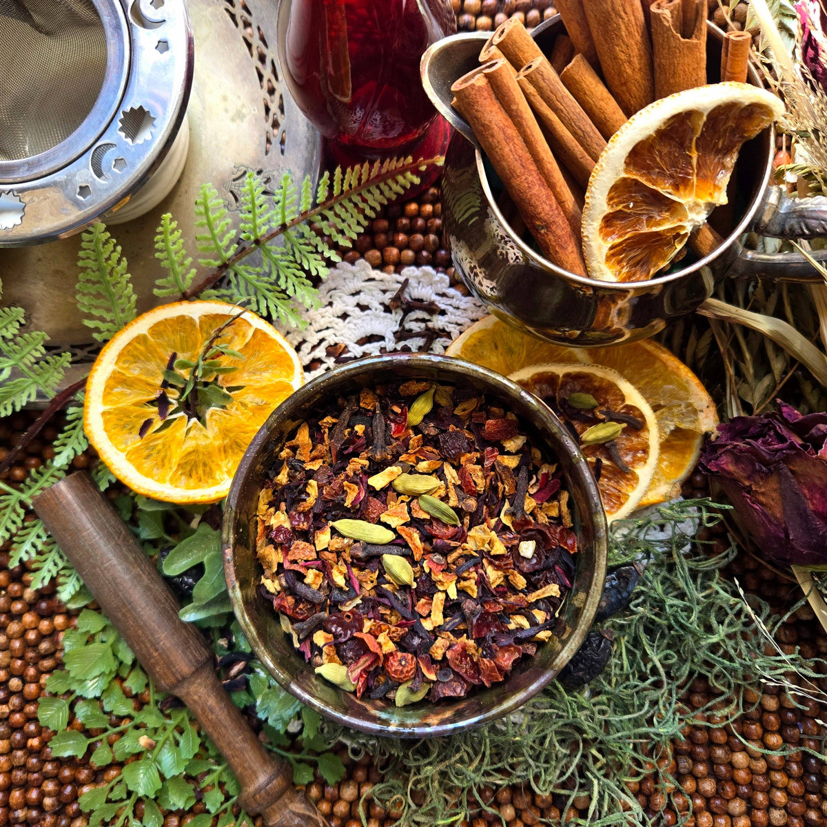 A bowl of loose-leaf herbal tea or spice blend surrounded by cinnamon sticks, dried orange slices, green herbs, and a wooden pestle in a styled flat-lay arrangement.