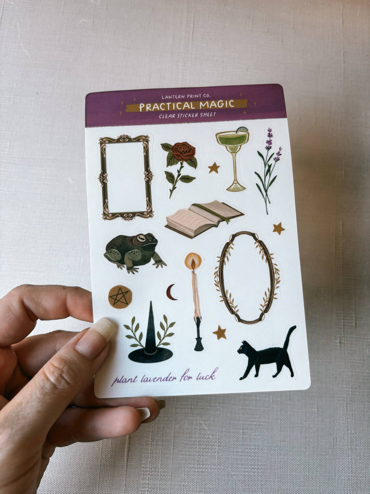 A hand holds a card of 'Practical Magic' cream sticker sheets by Lantern Print Co. The white sticker sheet features various mystical illustrations including a green frog, an open book, a martini glass, a black cat, and decorative frames.