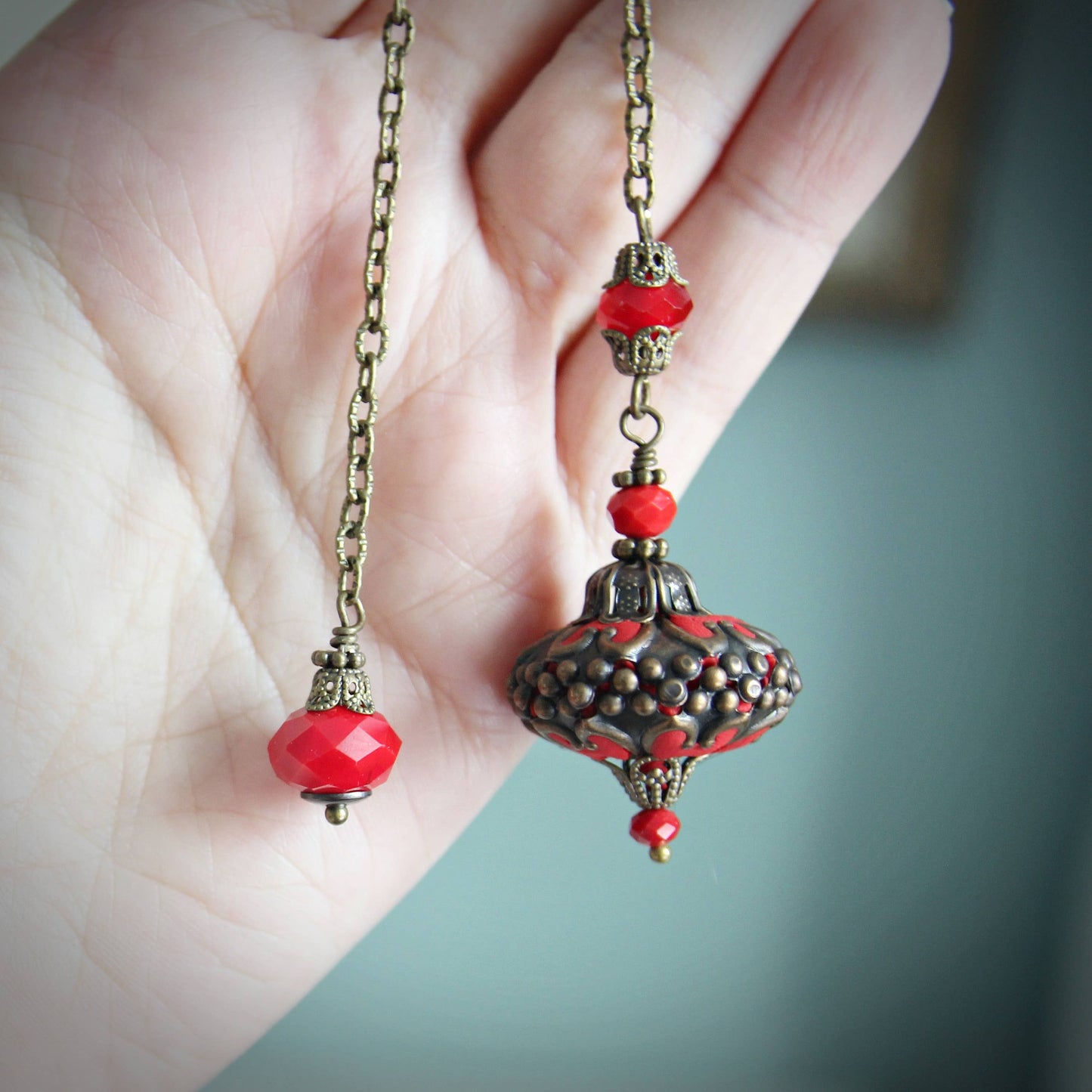 Decorative pendulum with red beads and intricate design held by a hand against a blurred background