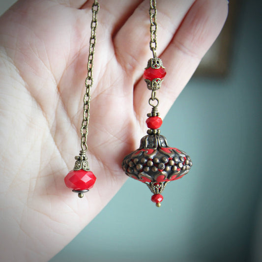 Decorative pendulum with red beads and intricate design held by a hand against a blurred background