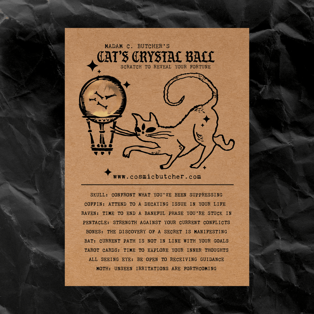 A rectangular kraft-paper style card with black printed text and line art reading 'Madam C. Butcher's Cat's Crystal Ball' and 'Scratch to reveal your fortune,' placed on a dark crumpled fabric background.