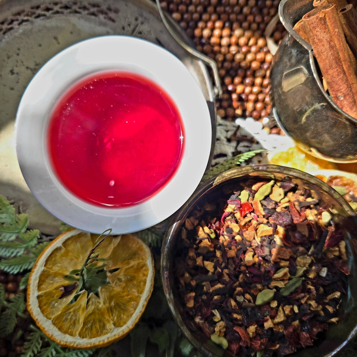 A top-down view of a vibrant red herbal tea mixture in a white cup, surrounded by its natural ingredients including a dried orange slice, whole spices, and cinnamon in a rustic setting.