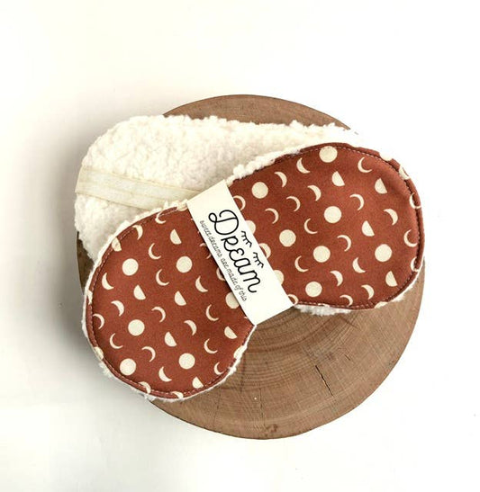 A pair of baby sleepers with a rust-brown and cream moon pattern, featuring a soft cream sherpa lining and a white paper band label that reads 'the Dream'. The sleepers are stacked on a round wooden slice against a plain white background.