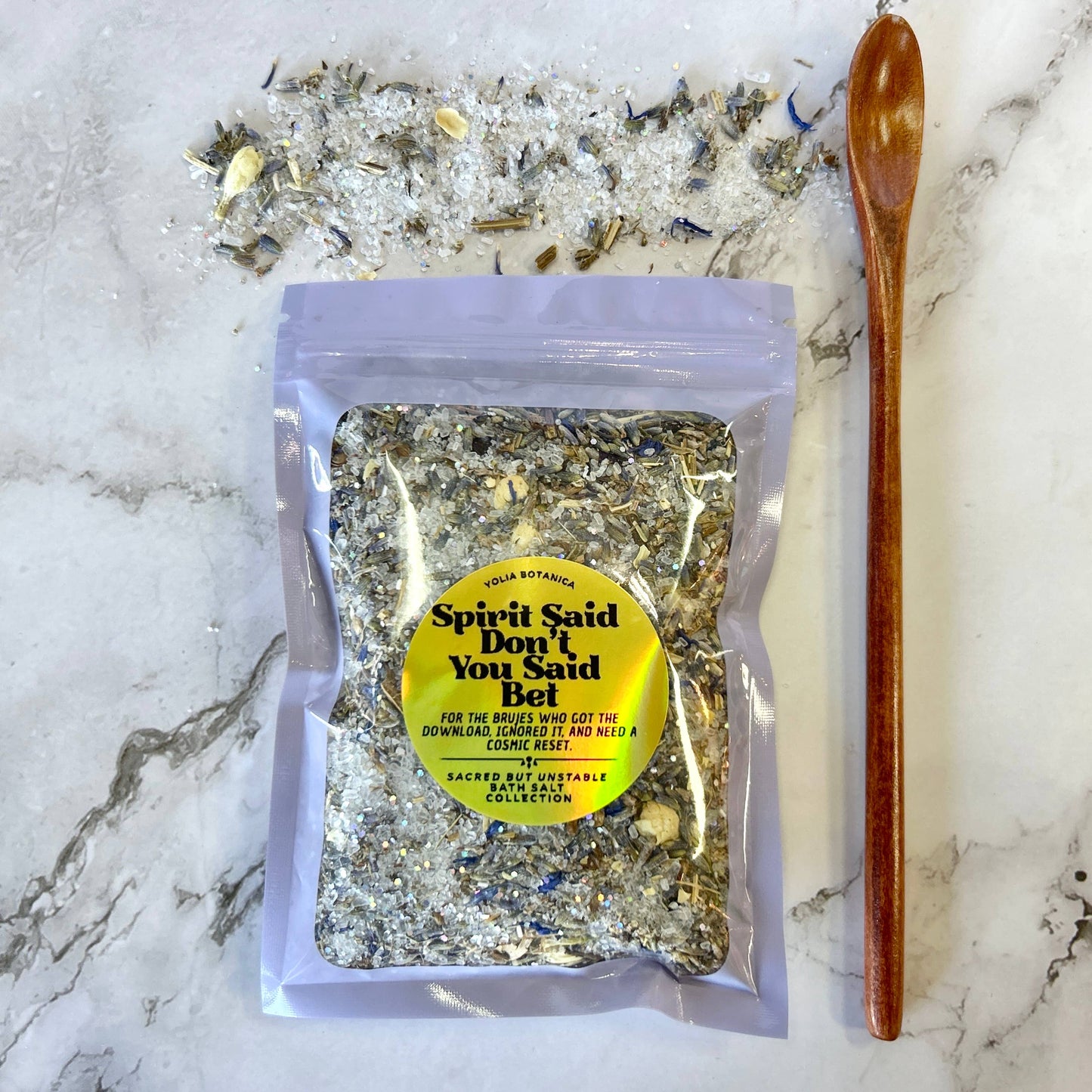A clear stand-up pouch containing a textured herbal salt blend, placed on a white marble surface with loose salt crystals scattered behind it and a wooden spoon to the right. The pouch has a yellow circular label reading 'Spirit Said Don't You Said Bel' and 'Sage Botanicals'.
