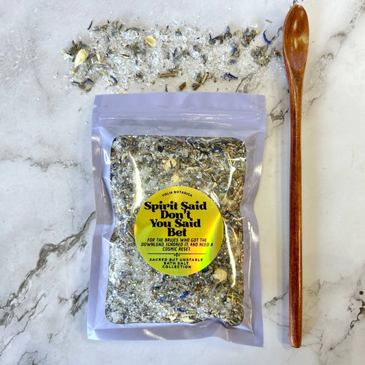 A clear stand-up pouch containing a textured herbal salt blend, placed on a white marble surface with loose salt crystals scattered behind it and a wooden spoon to the right. The pouch has a yellow circular label reading 'Spirit Said Don't You Said Bel' and 'Sage Botanicals'.