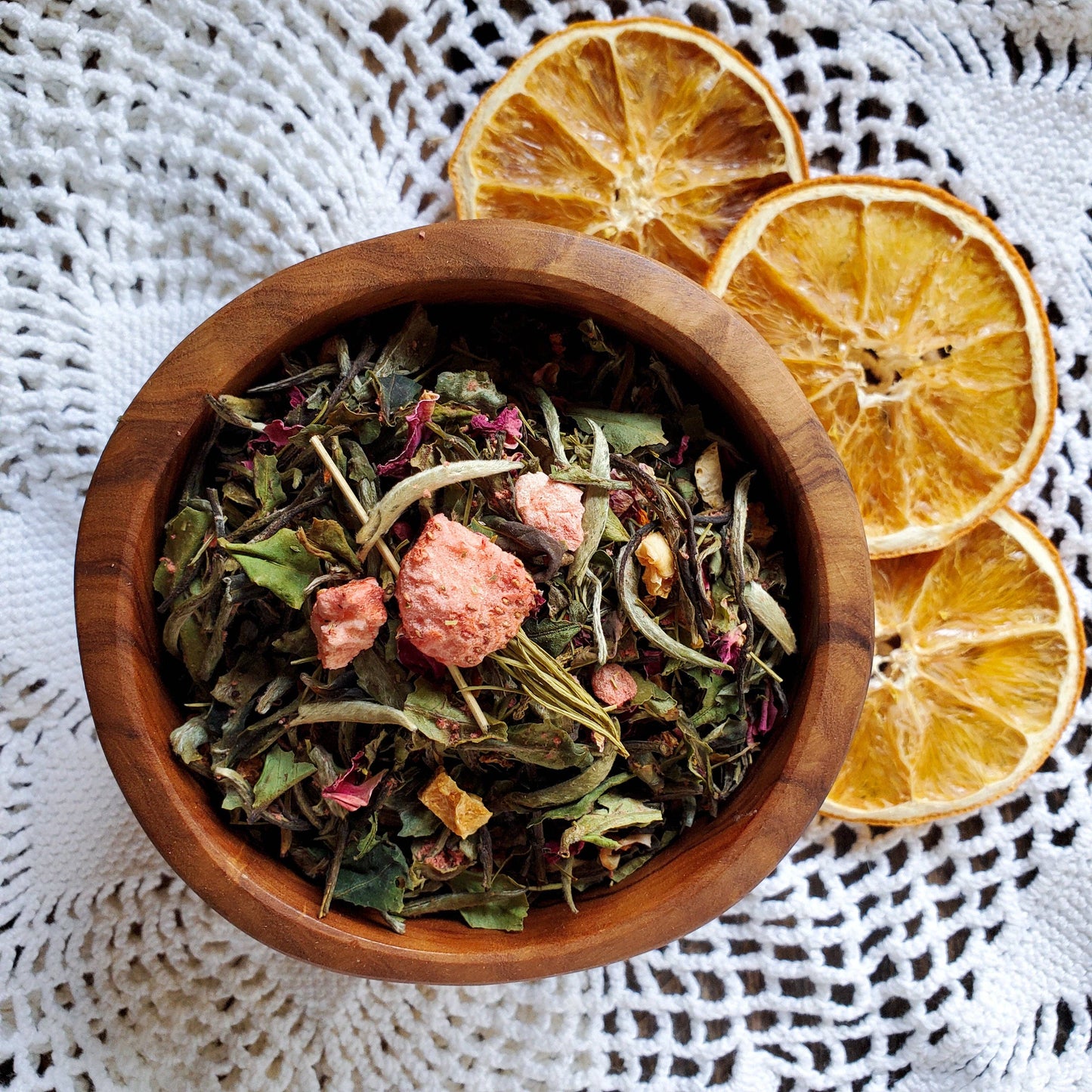 Wooden bowl of dried tea leaves with dried orange slices on a textured white surface