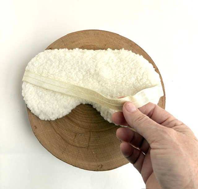 A white sherpa fleece sleep mask with a beige elastic strap, shown on a round wooden slice with a hand adjusting the strap.