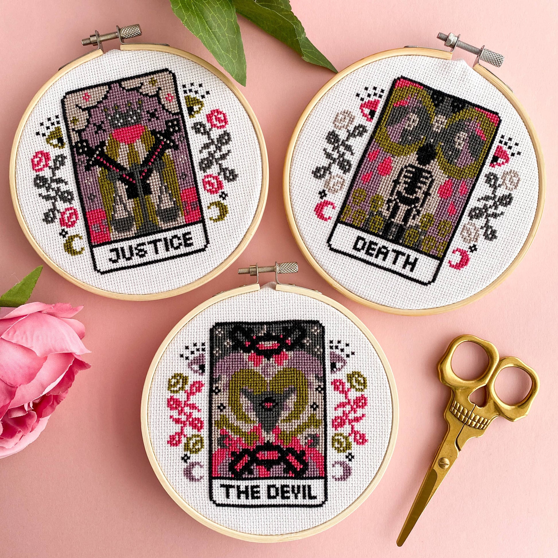 Three tarot card cross stitch pieces labeled 'Justice', 'Death', and 'The Devil' on a pink background with scissors and flowers.