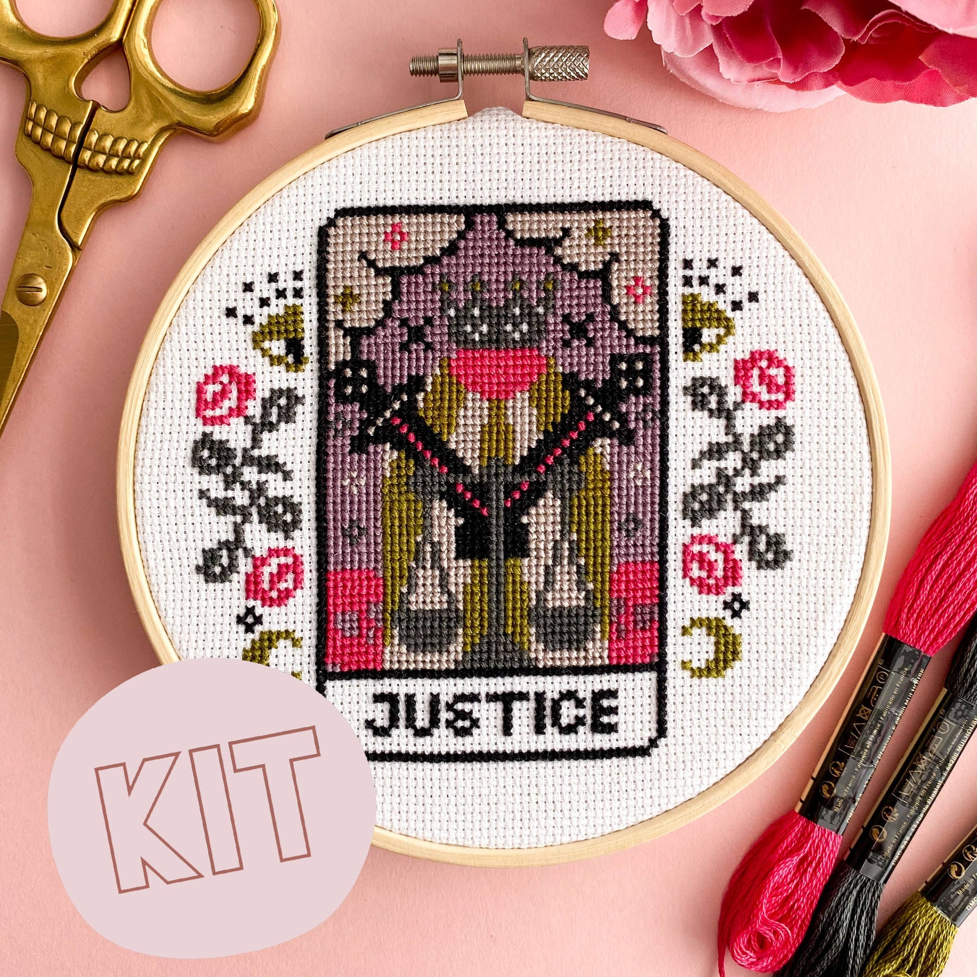 Cross-stitched tarot card design in an embroidery hoop with scissors and thread on a pink background.