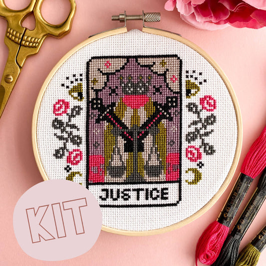 Cross-stitched tarot card design in an embroidery hoop with scissors and thread on a pink background.