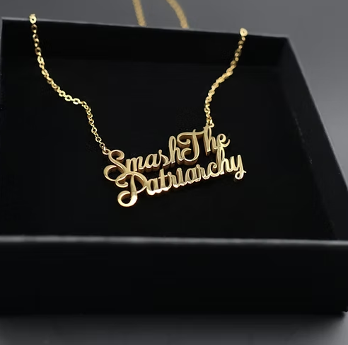 Close up of a gold Smash the Patriarchy Necklace on a black jewelry box.