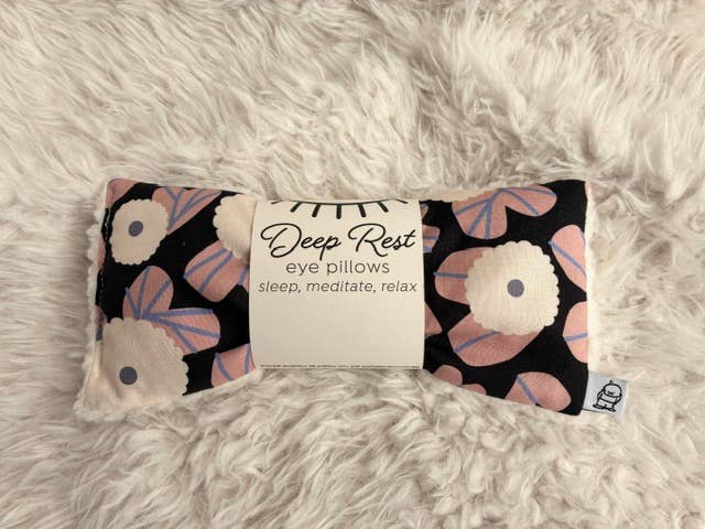 A rectangular eye pillow with a black background and a floral pattern in pink, beige, and blue, wrapped in a white paper band that reads 'Deep Rest eye pillows sleep, meditate, relax'.