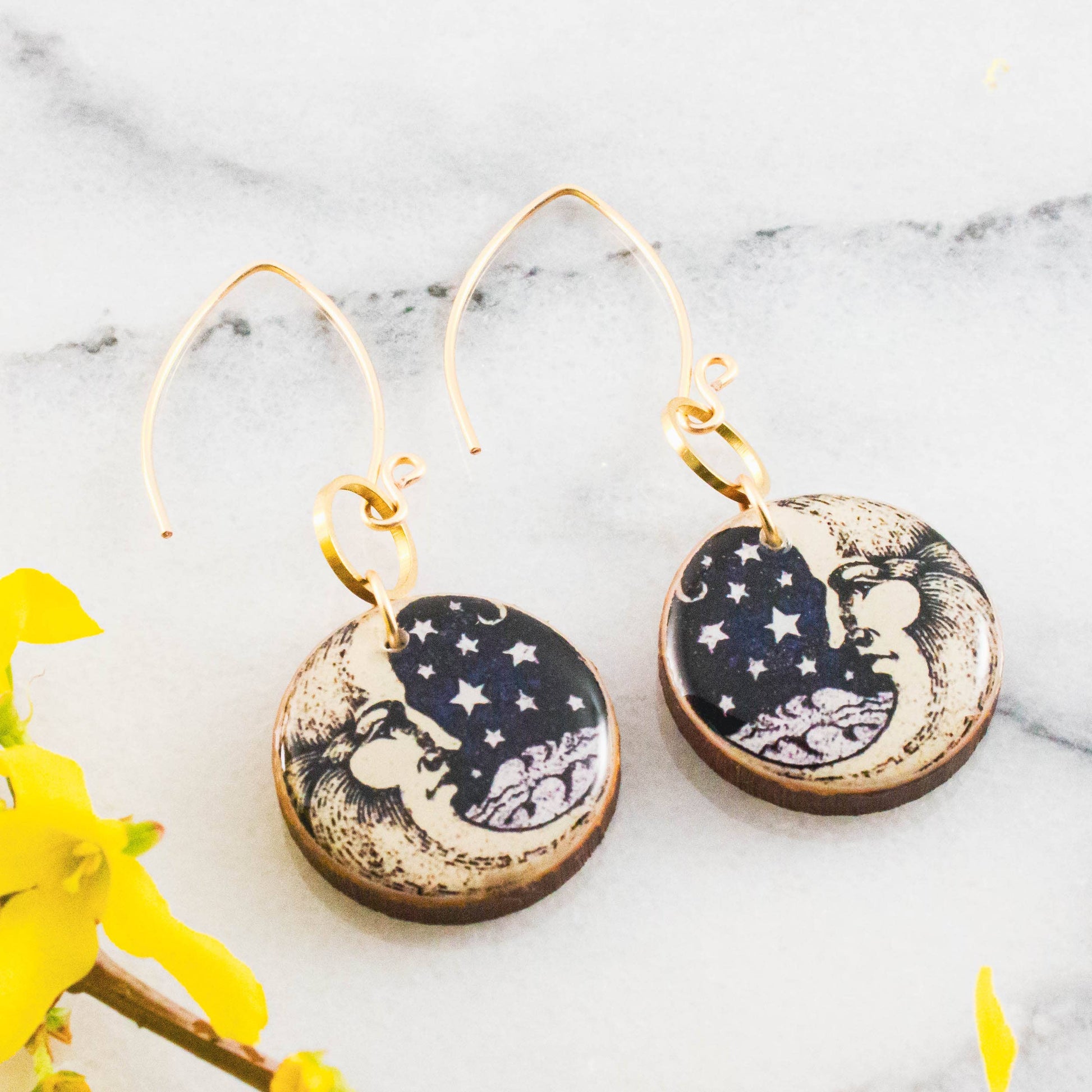 Pair of earrings with celestial design on a marble surface with yellow flowers.