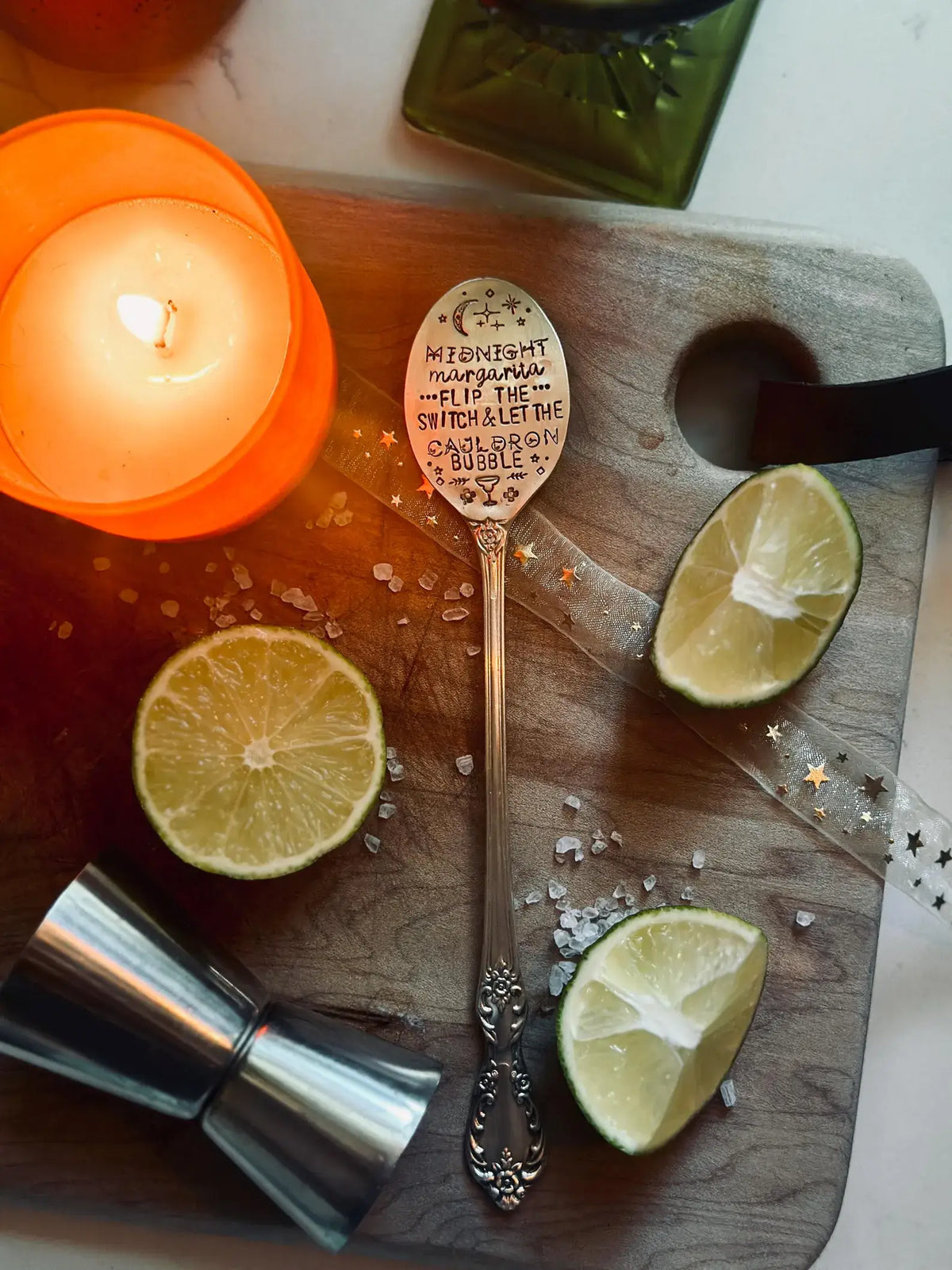 An ornate silver spoon with a hand-painted whimsical quote on the bowl, styled on a wooden cutting board with lime slices, a lit orange candle, a metal jigger, and coarse salt.