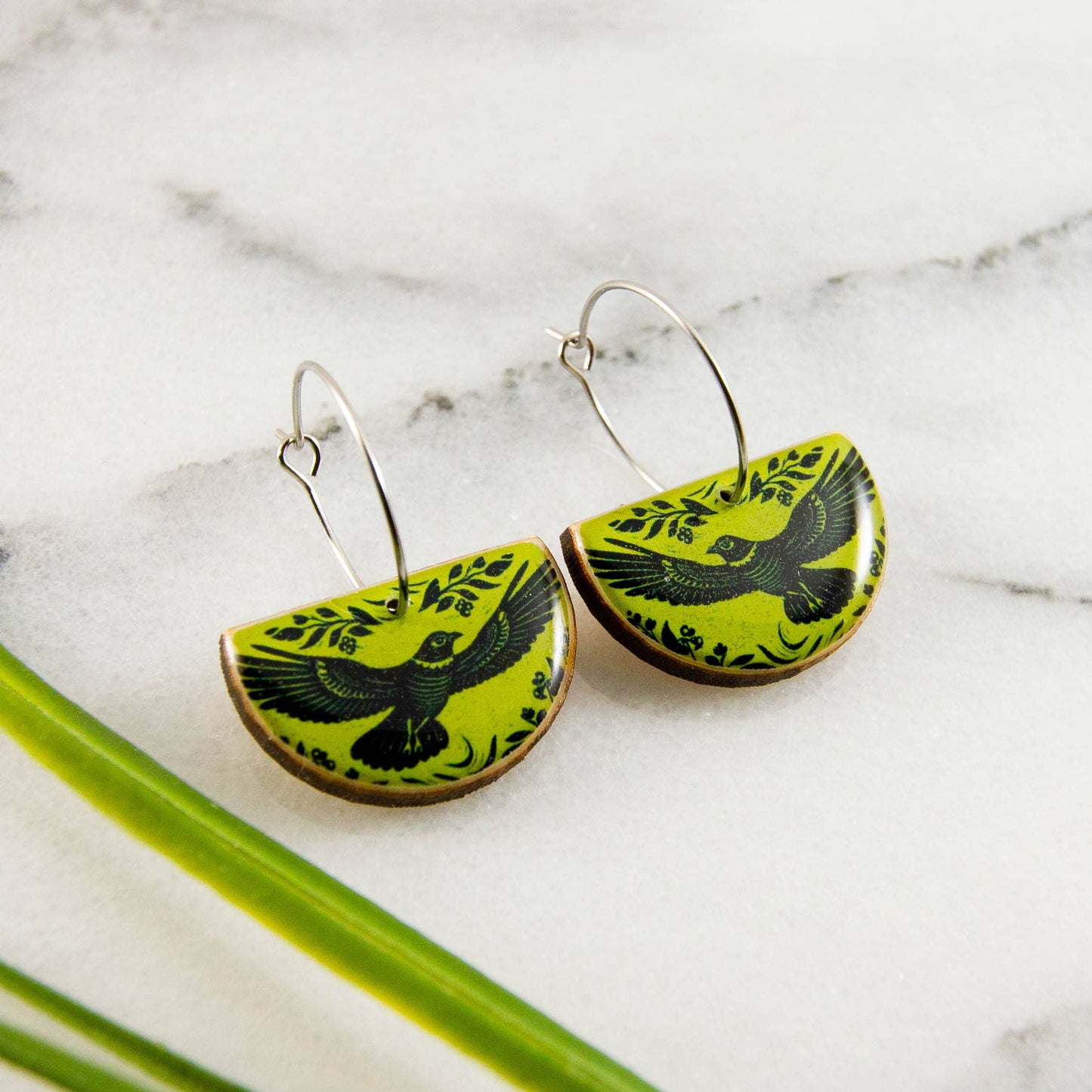 Green and black bird-patterned earrings on a marble surface with green leaves.