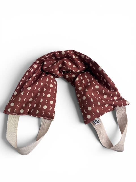 A baby carrier pillow in a dark reddish-brown fabric with a cream moon phase pattern, featuring beige shoulder straps and a small brand tag that reads '100%'.