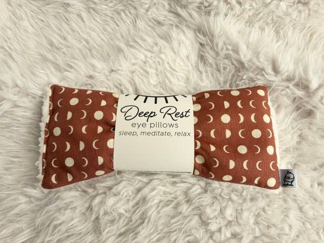 A Deep Rest eye pillow with a rust-brown and cream moon pattern, wrapped in a white paper band that reads 'Deep Rest eye pillows sleep, meditate, relax', placed on a white faux fur surface.