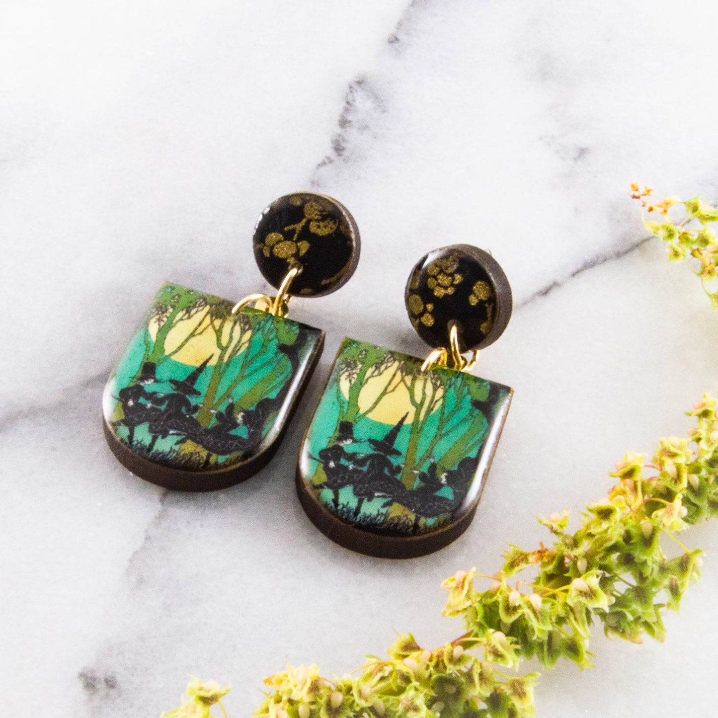Pair of earrings with nature-themed design on a marble surface with green leaves.
