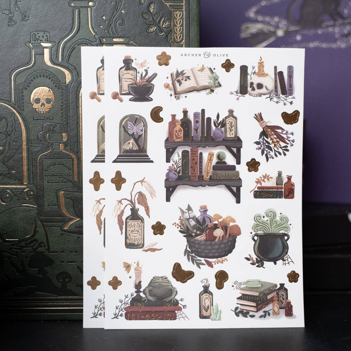 A sheet of decorative stickers featuring witch and potion-themed illustrations, including cauldrons, bottles, books, mushrooms, and butterflies, displayed in front of a dark patterned box.
