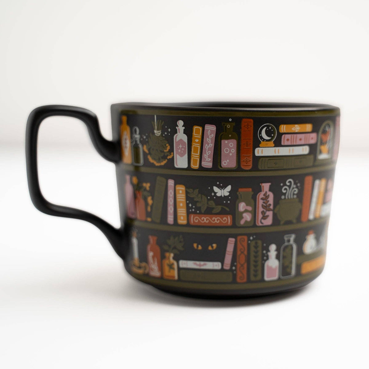 A black ceramic mug featuring a detailed illustration of shelves filled with books, potion bottles, and magical symbols in muted orange, pink, and white tones.