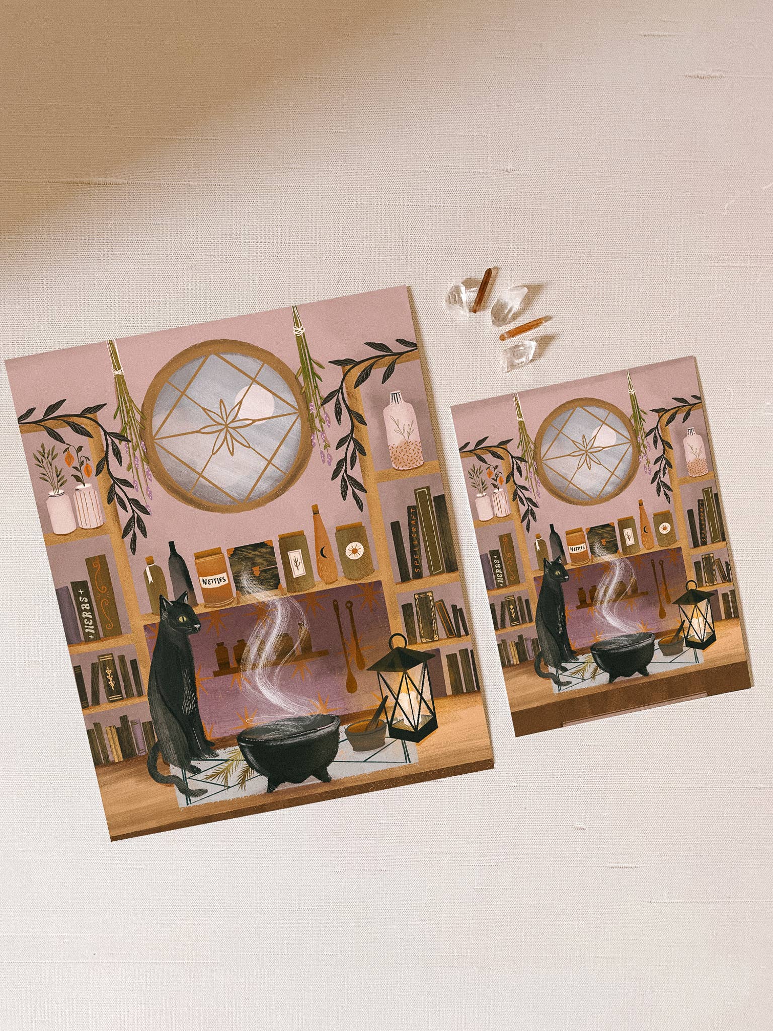 Two identical posters of a witch's kitchen scene with a cat, cauldron, and books on a pink background.