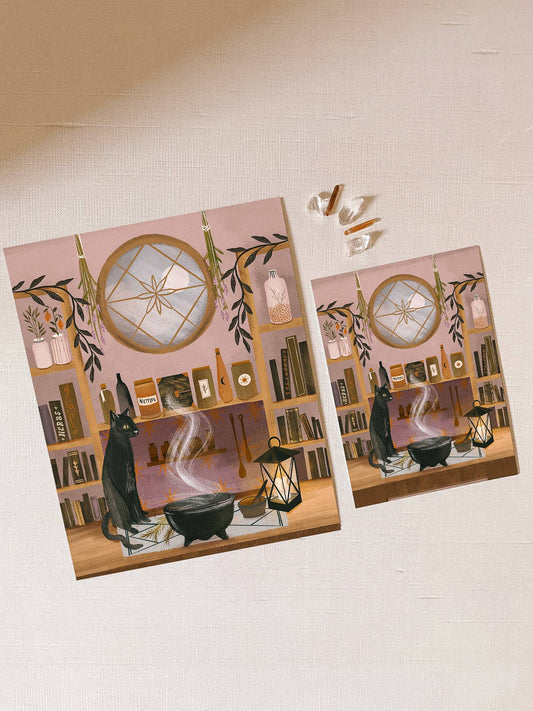 Two identical posters of a witch's kitchen scene with a cat, cauldron, and books on a pink background.