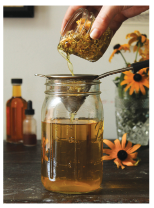 Hand pouring a liquid from a jar into a glass container with a strainer, surrounded by bottles and flowers.