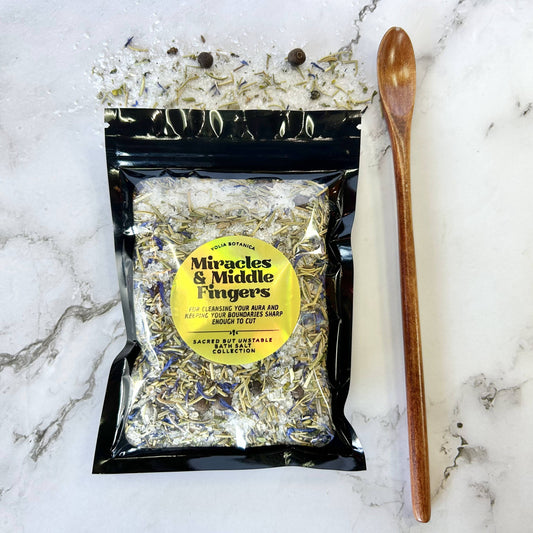 A black resealable pouch of dried herbs and spices labeled 'Miracles & Middle Fingers' by Sacred Spice Collection, placed on a white marble surface next to a wooden spoon.